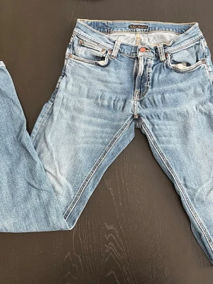 Ljusblå jeans från Nudie Jeans - Säljer ett par ljusblå jeans från Nudie Jeans med klassisk femficksdesign. Jeansen har kontrastsömmar och Nudie-loggan broderad på bakfickorna. Perfekt passform och tillverkade i mjukt denimtyg. OBS! Jeansen är ljusare på bild än verklighet, blivit strul med kameran som gjorde att Jeansen ser ljusare ut. Storleken på jeansen är W31 och L34