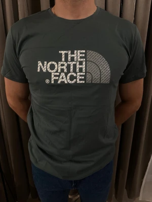 Mörkgrön t-shirt från The North Face - Snygg mörkgrön t-shirt från The North Face med stort logotryck i vitt och grått på bröstet. Trycket har ett mönster med texten 'The North Face' upprepat. T-shirten har rund hals och korta ärmar, perfekt för en chill och sportig look.