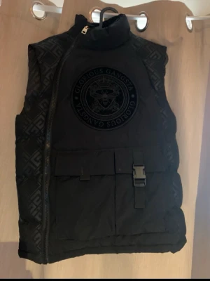 Svart dunväst från Glorious Gangsta - Säljer en svart dunväst från Glorious Gangsta med stort emblem på bröstet och mönstrade paneler på sidorna. Västen har en sned dragkedja, stor ficka med lock och spänne framtill. Perfekt för dig som vill ha en streetstyle-look.