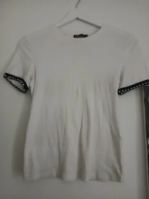 Vit t-shirt med pärldetaljer från Zara - Vit t-shirt från Zara i bomull med korta ärmar. Ärmsluten har svarta band med pärlor som ger en unik och snygg touch. Rund halsringning och rak passform. Perfekt för dig som gillar enkla plagg med coola detaljer. Det har tyvärr lostnat någon pärla och något märke på men välldigt fin💛