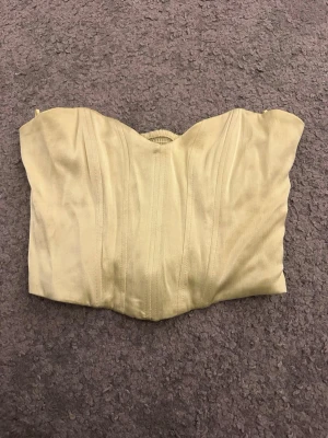 Ljusgul korsettopp från Zara XS - Säljer en ljusgul korsettopp från Zara i storlek XS. Toppen är axelbandslös med en snygg formad överdel och har smockad rygg för extra komfort. Materialet är mjukt och har en lätt glansig finish, med dragkedja i sidan för enkel på- och avtagning.