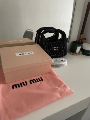 Svart quiltad handväska från Miu Miu - Säljer en svart quiltad handväska från Miu Miu med guldfärgade detaljer och logotyp framtill. Väskan är tillverkad i skinn och har ett snyggt mönster med dragkedja upptill. Medföljer axelrem och dustbag i rosa. Perfekt för dig som gillar lyxiga accessoarer.