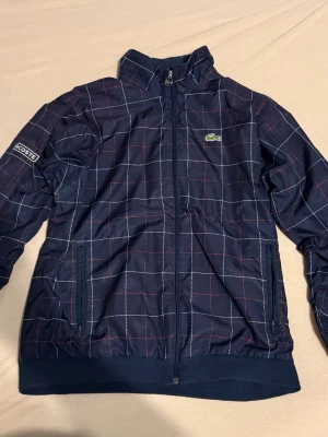 Mörkblå rutig vindjacka från Lacoste - Snygg mörkblå vindjacka från Lacoste med rutig design i vitt och rött. Jackan har dragkedja framtill, ståkrage och två sidofickor. Materialet är polyester och jackan har långa ärmar med muddar. Perfekt för dig som gillar sportig och clean stil.