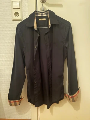 Svart Burberry skjorta S - Svart skjorta från Burberry med klassiskt rutigt mönster i beige och rött på insidan av krage och en diskret broderad logga på bröstet. Snygg och stilren design som sticker ut med de rutiga detaljerna. Skjortan är i mycket bra skick och bara använd 2 gånger.