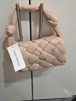 Beige quiltad axelväska Carin Wester - Säljer en beige axelväska från Carin Wester med quiltad design och bred, vadderad axelrem med knutdetalj. Väskan har magnetlås och flera fack inuti för smart organisering. Perfekt storlek för det viktigaste och riktigt snygg att bära över axeln.