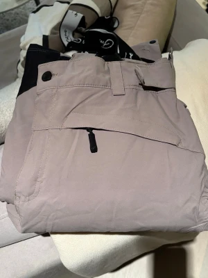 Beige täckbyxor med hängslen strl 40 - Säljer ett par beige täckbyxor från Five i storlek 40. Byxorna har svarta detaljer, justerbara hängslen med logga, justerbara i midjan, dragkedjor vid fickorna och förstärkning vid bensluten. Perfekta för vinterns alla äventyr i backen eller snön. Vind och vattentäta. Färgen på första bilden mest representativ.