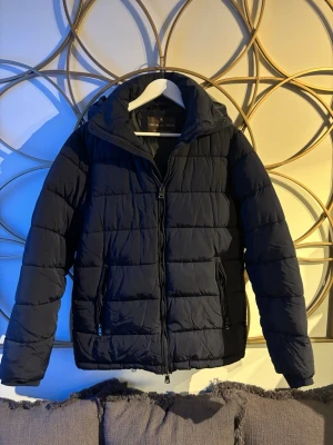 Mörkblå pufferjacka från Oscar Jacobson - Säljer en mörkblå pufferjacka från Oscar Jacobson med huva och dragkedja. Jackan är vattenavvisande och isolerande, perfekt för kalla dagar. Materialet är syntet och polyester, med quiltad design och två fickor med dragkedja framtill. Ny pris är 6000