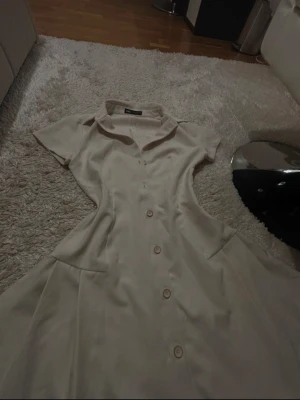 Beige kort klänning från Zara - Superfin beige kort klänning från Zara med knappar framtill och krage. Klänningen har korta ärmar och markerad midja med lätt utställd kjol. Perfekt för dig som gillar en stilren och fräsch look.