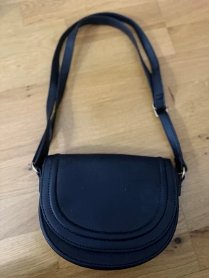 Svart axelväska i skinn - Snygg svart axelväska med rundad form och justerbar rem. Väskan har stilrena sömmar och är tillverkad i skinn, vilket ger en klassisk och tidlös look. Perfekt storlek för det viktigaste och enkel att matcha med olika outfits.