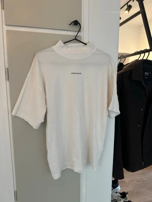 Vit t-shirt från Calvin Klein Jeans - Vit t-shirt från Calvin Klein Jeans med hög krage och diskret logga tryckt på bröstet. Modellen är kortärmad och har en loose passform som ger en chill vibe. Tillverkad i mjuk bomull som känns skön mot huden.