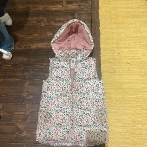 Blommig dunväst med huva - Säljer en söt blommig dunväst med huva och dragkedja framtill. Västen har en ljusrosa insida och små blommor i rosa, blått och grönt på utsidan. Perfekt för lager på lager under kyliga dagar.