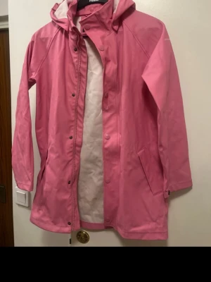 Rosa regnjacka med huva - Säljer en snygg rosa regnjacka från Didriksons med huva och långa ärmar. Jackan har dragkedja och knappar framtill samt två fickor på sidorna. Perfekt för regniga dagar och har en stilren look som sticker ut.