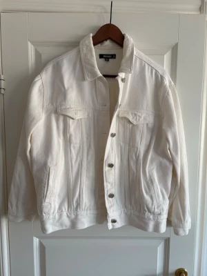 Vit jeansjacka från Missguided 40 - Säljer en fräsch vit jeansjacka från Missguided i klassisk modell med knappar framtill, två bröstfickor med lock och sidofickor. Jackan har lång ärm och en rak, lite loose passform. Perfekt att slänga över en hoodie eller t-shirt för en chill look.