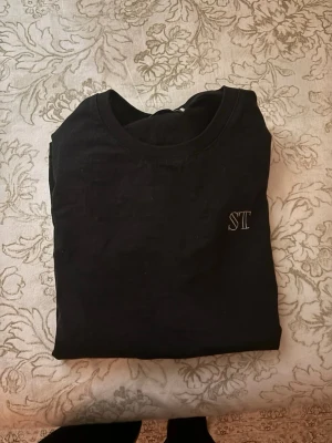 Svart t-shirt från St. Levitica - Svart t-shirt från St. Levitica med vit broderad logga på bröstet och stor text på ryggen. Klassisk rund hals och korta ärmar. Materialet är mjuk bomull som känns skönt mot huden. Perfekt för dig som gillar streetwear med tydliga detaljer.