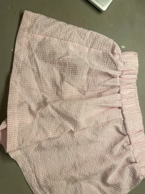 Rosa och vita randiga shorts - Säljer ett par ljusrosa och vita shorts med randigt mönster och elastisk midja. De har en loose passform och är tillverkade i ett mjukt bomullstyg med struktur. Framtill finns två små vita knappar som detalj. Perfekta för varma dagar!