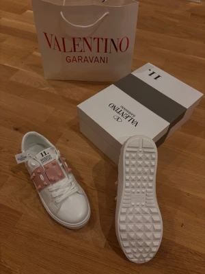 Valentino Rockstud Untitled sneakers - Säljer ett par vita Valentino Rockstud Untitled sneakers med ljusrosa band och ikoniska pyramidnitar. Skorna har snörning, rund tå och platt sula. Tillverkade i skinn med detaljer i rosa och vita toner. Kommer med originalkartong och påse.