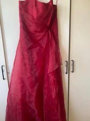 Röd balklänning  - Säljer en elegant röd långklänning i ett skirt och glansigt material. Klänningen har smala axelband, vackra volangdetaljer på sidan och en dekorativ pärlspänne i midjan. Baksidan har snygg snörning som ger en cool touch och bra passform.