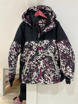Leopardmönstrad vinterjacka Wall Ride - Snygg vinterjacka från Wall Ride med leopardmönster i svart, vit och lila samt detaljer i orange. Jackan har huva, dragkedja, reflexdetaljer och justerbara muddar. Perfekt för dig som vill sticka ut med cool stil under kalla dagar.