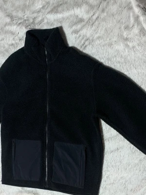 Svart teddyjacka med dragkedja - Mysig svart teddyjacka med hög krage och hel dragkedja framtill. Jackan har två stora fickor i nylonmaterial på framsidan som ger en cool kontrast. Perfekt för att hålla värmen under kyliga dagar och har en enkel, stilren look.