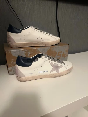 Golden Goose Superstar sneakers vit/grå - Golden Goose Superstar sneakers i vitt skinn med grå mockadetaljer och grå snörning. Skorna har en ikonisk stjärna på sidan, mörkblå hälkappa och vintage-inspirerad sula. Perfekta för dig som gillar en cool och avslappnad stil.