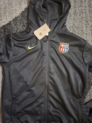 Nike FC Barcelona träningsset svart XL - Säljer ett svart Nike FC Barcelona träningsset i storlek XL. Setet består av en hoodie med dragkedja och byxor, båda med guldiga Nike-loggor och Barcelonas klubbmärke. Tillverkat i mjukt, syntetiskt material som passar perfekt till träning.