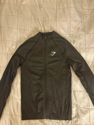 Svart zip-tröja från Gymshark - Säljer en helt oanvänd svart långärmad zip-tröja från Gymshark. Lappen är bortklippt men aldrig använd. Tröjan har en dragkedja framtill och ett vitt Gymshark-logo på bröstet, perfekt för träning eller chill. Nypris 550 kr. 