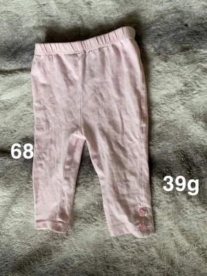 Ljusrosa leggings med knappar, stl 68 - Söta ljusrosa leggings med resår i midjan. Nedtill på ena benet finns två små dekorativa knappar i samma färg som byxorna. Perfekta för en bekväm och gullig vardagslook till de minsta.🌸 Fint använt skick. Storlek 68