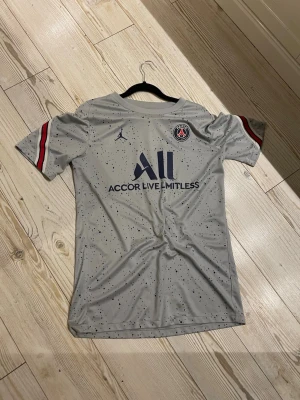 Grå PSG x Jordan fotbollströja M - Säljer en grå PSG x Jordan fotbollströja i slim fit med mörkblå och röda detaljer på ärmarna. Tröjan har ett prickigt mönster och Paris Saint-Germain-logga på bröstet. Materialet är syntet och perfekt för fotboll. Snygg design med Accor Live Limitless-tryck.