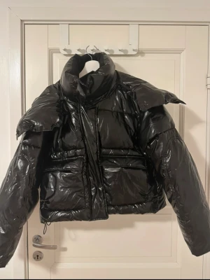 Svart pufferjacka från Calvin Klein Jeans - Säljer en svart, glansig pufferjacka från Calvin Klein Jeans med hög krage och croppad passform. Jackan har två stora fickor framtill och dragkedja. Materialet är syntet och polyester, perfekt för kalla dagar. Snygg och trendig look med oversized känsla. 