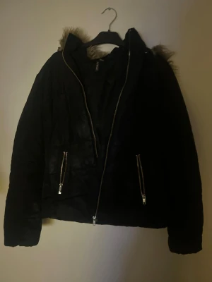 Svart pufferjacka med fuskpäls från H&M - Svart pufferjacka från H&M med fuskpäls på huvan och två fickor med dragkedja. Jackan har en snygg, lite boxig passform och är tillverkad i polyester. Perfekt för kalla dagar när du vill vara både varm och stilren.