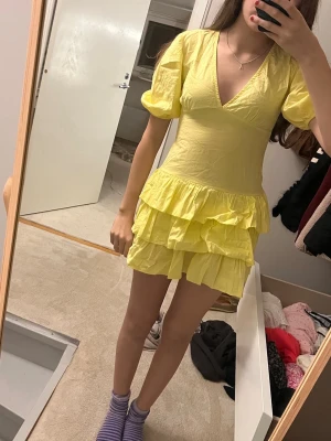 Gul kort volangklänning med puffärm - Supersöt gul klänning med volanger och puffiga korta ärmar. Klänningen har en djup v-ringning och flera lager volanger på kjolen som ger en lekfull look. Perfekt för dig som gillar färg och vill sticka ut. Materialet känns lätt och luftigt. Kp vid behov av mer bilder!