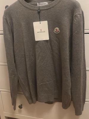 Grå stickad tröja från Moncler - Snygg grå stickad tröja från Moncler med rund hals och ribbade muddar. Tröjan har Moncler-logga på bröstet och är långärmad, perfekt för dig som gillar stilrena och klassiska plagg. Passar dig som vill ha en enkel men exklusiv look.