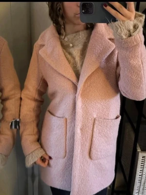 Ljusrosa kappa från ICHI - Superfin ljusrosa kappa från ICHI med stora fickor framtill och klassisk krage. Jackan är i ett mjukt, ulligt material och har en rak passform som känns både chill och stilren. Perfekt för dig som vill ha en mysig men snygg look under höst och vinter.