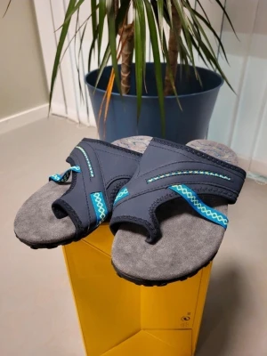 Grå och marinblå sandaler med mönster 38 - Säljer ett par sandaler i grått och marinblått med coola blå och gula detaljer. Ovandelen är i syntetmaterial med broderade mönster och mjuk innersula i mocka-look. Sulan är svart med bladliknande struktur för extra grepp. Perfekt för sommaren!