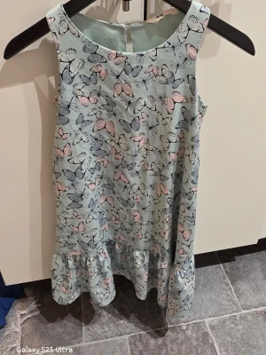 Ljusblå kortklänning med fjärilar H&M - Supersöt ljusblå kortklänning från H&M med ett gulligt fjärilsmönster i rosa och grå toner. Klänningen är ärmlös och har en volang längst ner. Tillverkad i mjuk och lätt polyester som känns skön mot huden.