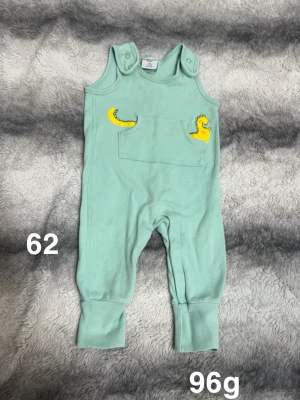 Mintgrön byxdress med dinosauriedetaljer - Supergullig mintgrön byxdress för baby med tryckta gula dinosauriesvansar på fickan framtill. praktiska knappar på axlarna för enkel påklädning. Perfekt för små äventyr och lek.🌸 Fint använt skick. Storlek 62