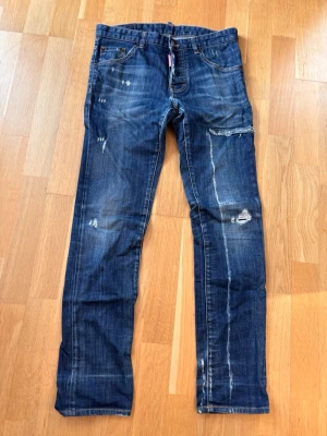 Blå jeans från Dsquared2 med slitningar - Säljer ett par blå jeans från Dsquared2 med coola slitningar och detaljerade sömmar. Jeansen har klassisk femficksdesign, raka ben och en mörk tvätt med ljusare partier. Materialet är robust denim och passformen är normal. Perfekt för dig som gillar streetstyle. OBS: lagade/förstärkta i grenen enl bild.