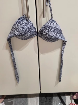 Leopardmönstrad bikiniöverdel Andy M - Snygg bikiniöverdel från Andy by Frank Dandy i storlek M. Den har ett grått och svart leopardmönster, trekantsformade kupor och knytband i nacken och ryggen. Materialet är mjuk polyester med stretch för skön passform. Perfekt för strandhäng!