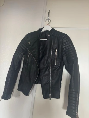 Svart skinnjacka från Voyelles - Snygg svart skinnjacka från Voyelles med coola quiltade detaljer på axlar och ärmar. Jackan har flera silvriga dragkedjor, bälten med spännen i midjan och en asymmetrisk front. Perfekt för dig som gillar edgy stil.