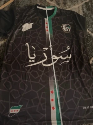 Syrien  - Säljer en svart fotbollströja med geometriskt mönster och gröna, vita samt röda detaljer. Tröjan har tryck med arabiska bokstäver och flera stjärnor på framsidan. Perfekt för dig som gillar unika matchtröjor.