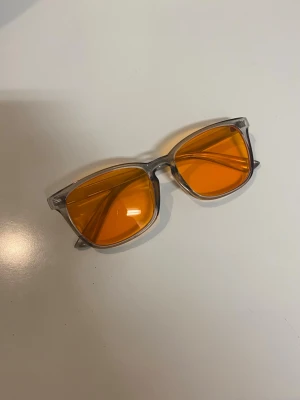 Blåljusglasögon - Ett helt nytt par glasögon med rektangulär form och orangefärgade glas. Bågarna är i en transparent grå färg. De används för att blockera blåljus inför sömn eller avslappnad. Från Aouhra.
