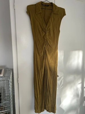 Olivgrön långklänning från Zara - Säljer en olivgrön långklänning från Zara med subtilt blommönster i tyget. Klänningen har v-ringning, krage och snygg rynkning framtill som ger en cool siluett. Ärmlös modell i mjukt, glansigt material som känns lyxigt och skönt mot huden. Den har inga anmärkningar men behöver strykas 