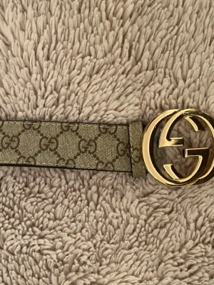 Guccee bälte beige/guld - Säljer ett klassiskt GG bälte i beige med det ikoniska GG-mönstret och stor guldfärgad logga som spänne. Bältet är gjort i skinn och har exklusiv känsla, perfekt för att lyfta vilken outfit som helst.