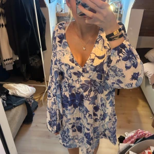 Blommig vit och blå kort mini klänning v ringad  - Obs! se hål som ej är synliga pga volang /fallet på klänningen , därav priset. Säljer en söt kortklänning med vit botten och blått blommigt mönster. Klänningen har v-ringad hals, långa puffärmar med resår vid ärmslut och en luftig passform. Perfekt för varma dagar och har en romantisk vibe. Materialet känns lätt och skönt mot huden.