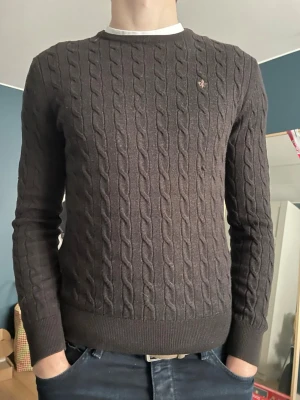 Mörkbrun kabelstickad tröja Morris - Säljer en mörkbrun kabelstickad tröja från Morris i 100% merinoull. Tröjan har rund hals, ribbade muddar och ett litet broderat emblem på bröstet. Perfekt för dig som gillar klassisk och stilren look.