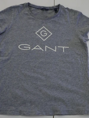 Grå t-shirt från Gant med logga - Snygg grå t-shirt från Gant med stor vit logga på bröstet. Klassisk rund halsringning och korta ärmar. Tillverkad i mjuk bomull som känns skön mot huden. Perfekt till jeans eller shorts för en clean och enkel look.
