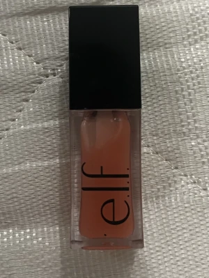 e.l.f. Glow Reviver Lip Oil - Säljer en e.l.f. Glow Reviver Lip Oil i en snygg persikofärgad ton. Kommer i en stilren, genomskinlig plastförpackning med svart lock. Perfekt för dig som vill ha glansiga och återfuktade läppar med en fräsch finish.