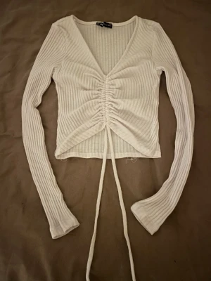 Vit ribbad omlottopp från RB Sister - Säljer en vit långärmad topp från RB Sister i storlek XS. Toppen är ribbad med omlottdesign och har snörning framtill som ger en snygg rynkad effekt. Materialet känns mjukt och stretchigt, perfekt för att styla med jeans eller kjol.