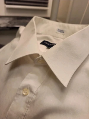 Vit skjorta från Hagenfeldt, storlek L - Vit skjorta från Hagenfeldt i storlek L med klassisk krage och bröstficka. Materialet är 55% bomull och 45% polyester, med diskret prickigt mönster i tyget. Skjortan har långa ärmar och knappar framtill samt vid manschetterna. Perfekt för dig som gillar stilrena och enkla plagg.