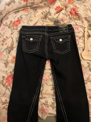 True Religion Joey Low Rise Flare jeans - Svarta jeans från True Religion, modell Joey Low Rise Flare. Snygga kontrastsömmar i vitt och röda detaljer, klassiska fickor med lock och silverfärgade knappar. Flare-ben och låg midja ger en cool siluett. Materialet är jeans med stretch för skön passform.